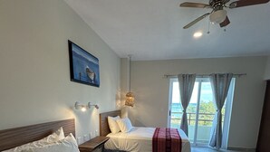 Quarto de hotel com duas camas, um ventilador de teto, um quadro na parede e vista pela janela.