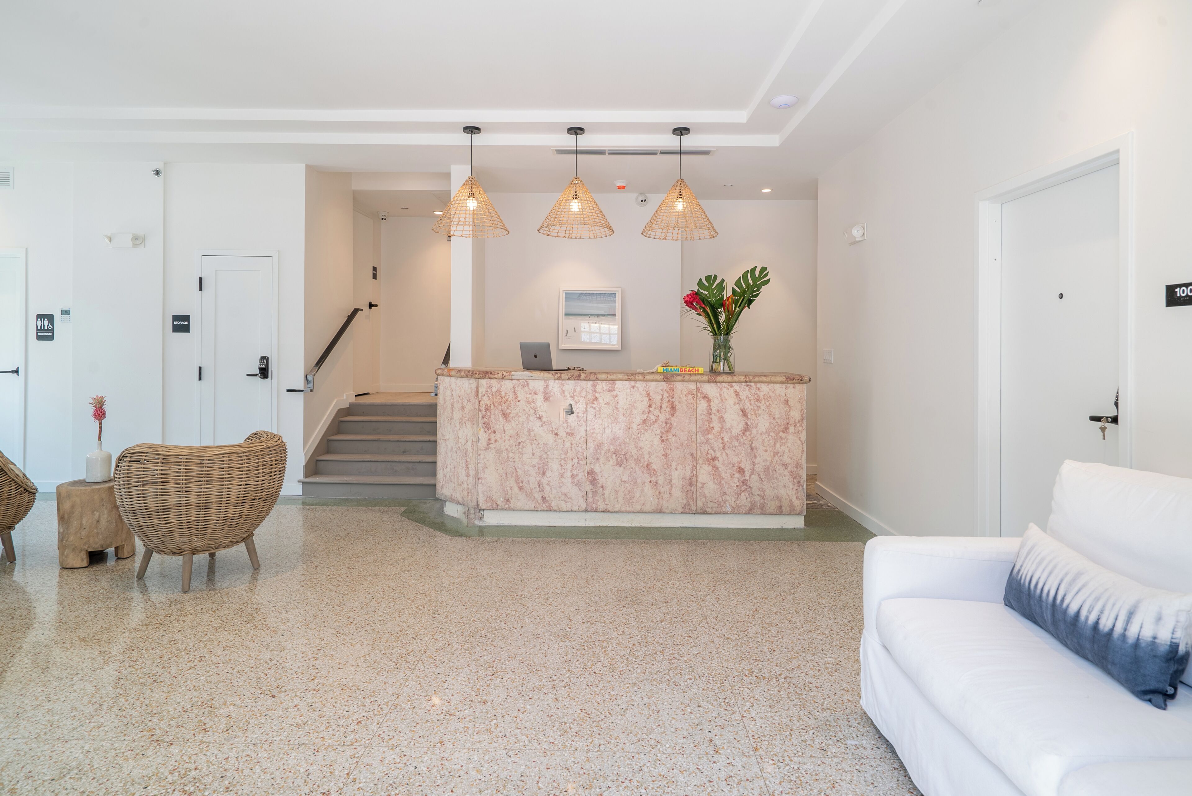 A reception area with a marble counter, wicker chairs, a white sofa, and a staircase leading to upper floors.