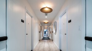 A hotel hallway with a light wood floor, white walls, and black door handles. There are numbered doors on either side, and a circular light fixture hangs from the ceiling. - Casa Ocean Queen Bed Front Room (Miami Beach)