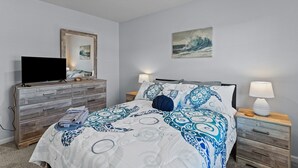 Un dormitorio con cama, cómoda, televisor, mesita de noche y un cuadro colgado en la pared.