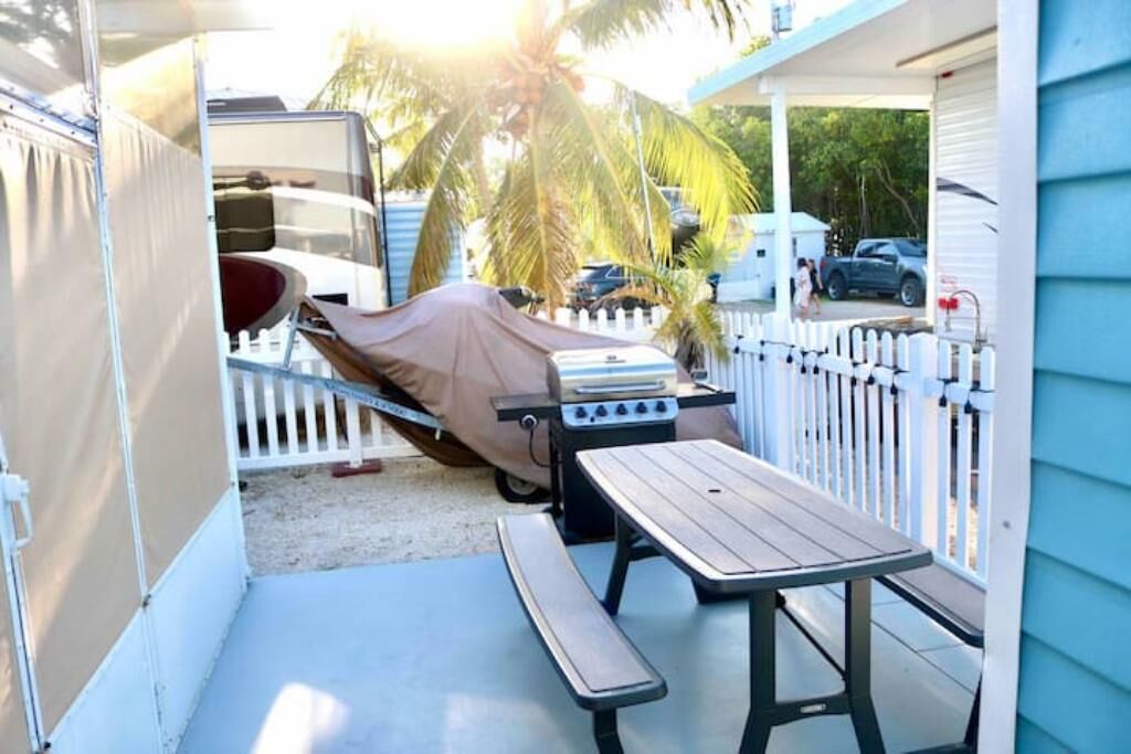 A covered patio with a grill and picnic table, surrounded by a white picket fence and palm trees.