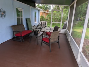 A covered patio with wicker furniture and red cushions, a bicycle, and a view of a green garden.