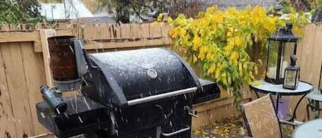 Una parrilla para barbacoa en una terraza de madera con un cubo de basura azul y una mesa con sillas.
