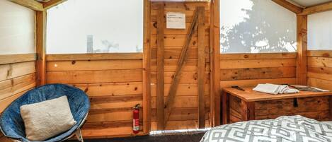 A wooden cabin room with a bed, a blue chair with a pillow, a small table, and a fire extinguisher.
