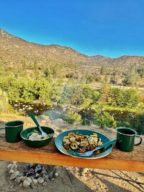 A meal setup with a plate of food, two green mugs, and a spoon on a wooden surface with a view of a river and mountains in the background. - SoCal Camping (Kernville)