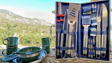 A camping gear set with utensils and cookware, including a knife, spatula, and pots, displayed on a wooden surface with a mountainous backdrop.
