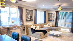A modern living room with a sofa, coffee table, and chairs. There is a dining area with a table and chairs. The room has large windows with curtains and artwork on the walls. - Peaceful 3bed3bath 2300sqft Home Centrally Located in Heart of Silicon Valley (Santa Clara)