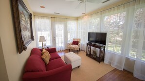 Una sala de estar con un sofá rojo, un otomano blanco, un televisor sobre un mueble de madera y una silla con cojín rojo.