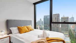 A modern hotel room with a large bed, a nightstand, and a view of the city skyline through a large window. - Stunning City Views 2B2B apt in CBD (Melbourne)