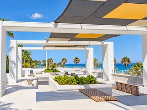 A covered outdoor seating area with benches and planters, overlooking a beach with palm trees and the ocean.