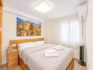 A bedroom with a bed, bedside table, lamp, air conditioning unit, and a painting on the wall.