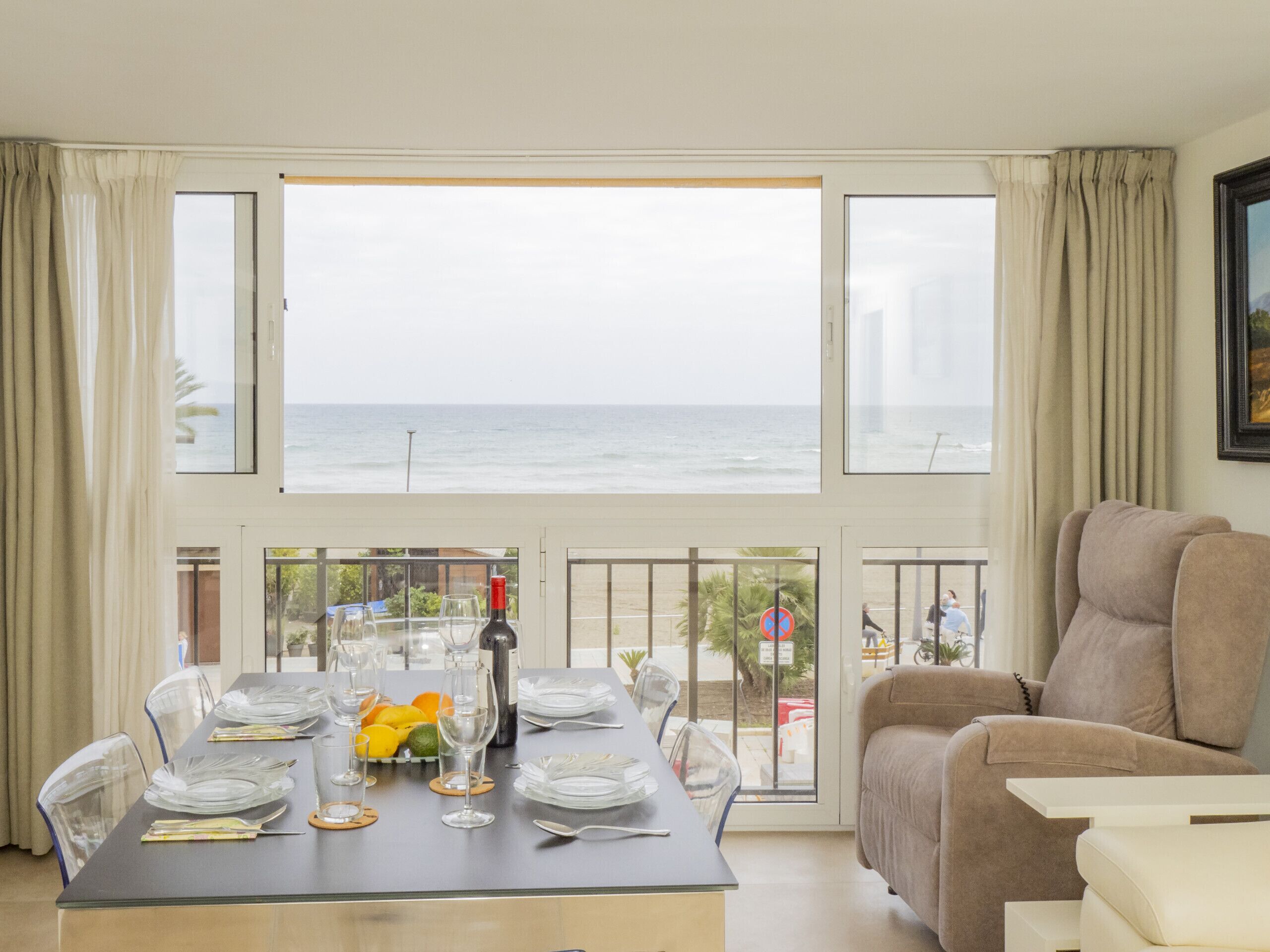 Un salón con una mesa de comedor dispuesta para seis personas, ventanas amplias con vista al mar y un sofá color beige.