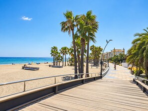 A wooden boardwalk leading to a sandy beach with palm trees, a clear blue sky, and a calm sea.