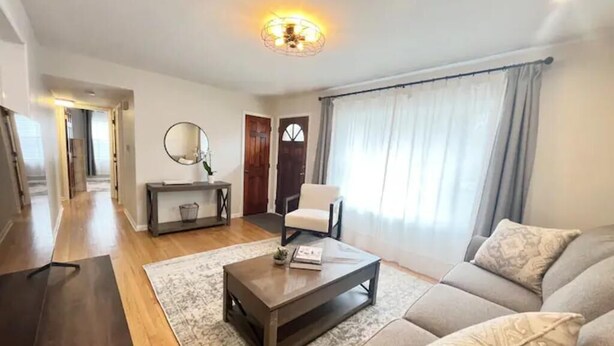 Uma sala de estar moderna com sofá, mesa de centro e espelho. Há uma porta que dá para outro quarto e uma janela com cortinas.