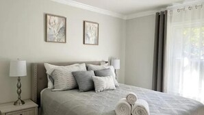 Uma cama bem arrumada com colcha acolchoada, duas pinturas penduradas na parede e uma janela com cortinas.