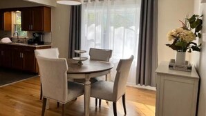 Uma sala de jantar com uma mesa redonda, quatro cadeiras, um lustre pendurado e vista pela janela com cortinas.
