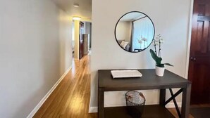 Um corredor com piso de madeira, um espelho redondo na parede, uma pequena mesa com bandeja branca e um vaso de planta.