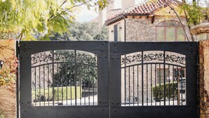 Un portail en fer forgé orné de motifs complexes, donnant accès à un bâtiment en pierre doté d’un toit en tuiles et d’une fenêtre bien visible.