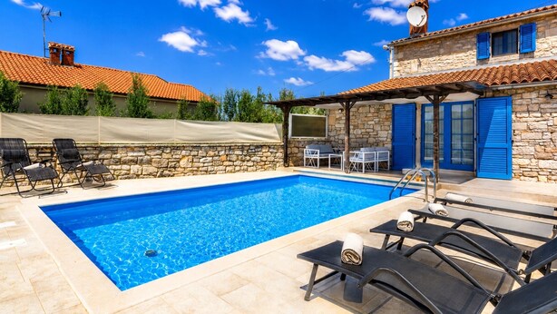 A swimming pool with lounge chairs, a stone building with blue shutters, and a clear blue sky.