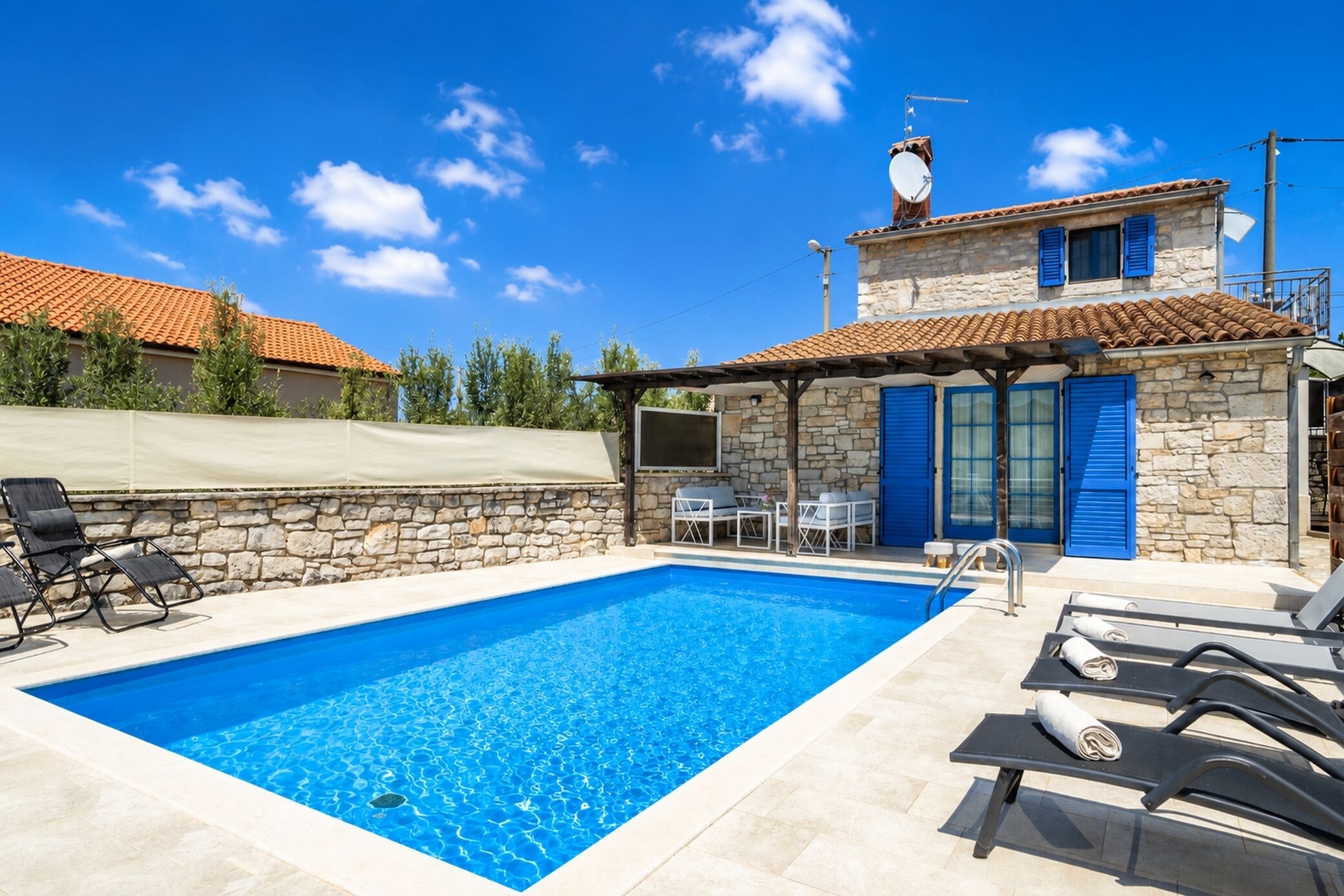 A swimming pool with lounge chairs, a stone building with blue shutters, and a clear blue sky.