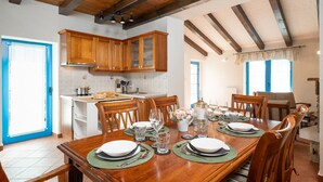 A dining area with a wooden table set for four, surrounded by wooden chairs. A kitchen with wooden cabinets and a blue door is visible in the background.