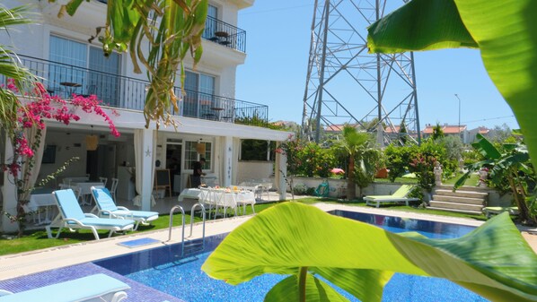 A pool area with lounge chairs, a dining table, and a building with balconies. - Lavinia Garden Alaçatı (Çeşme)