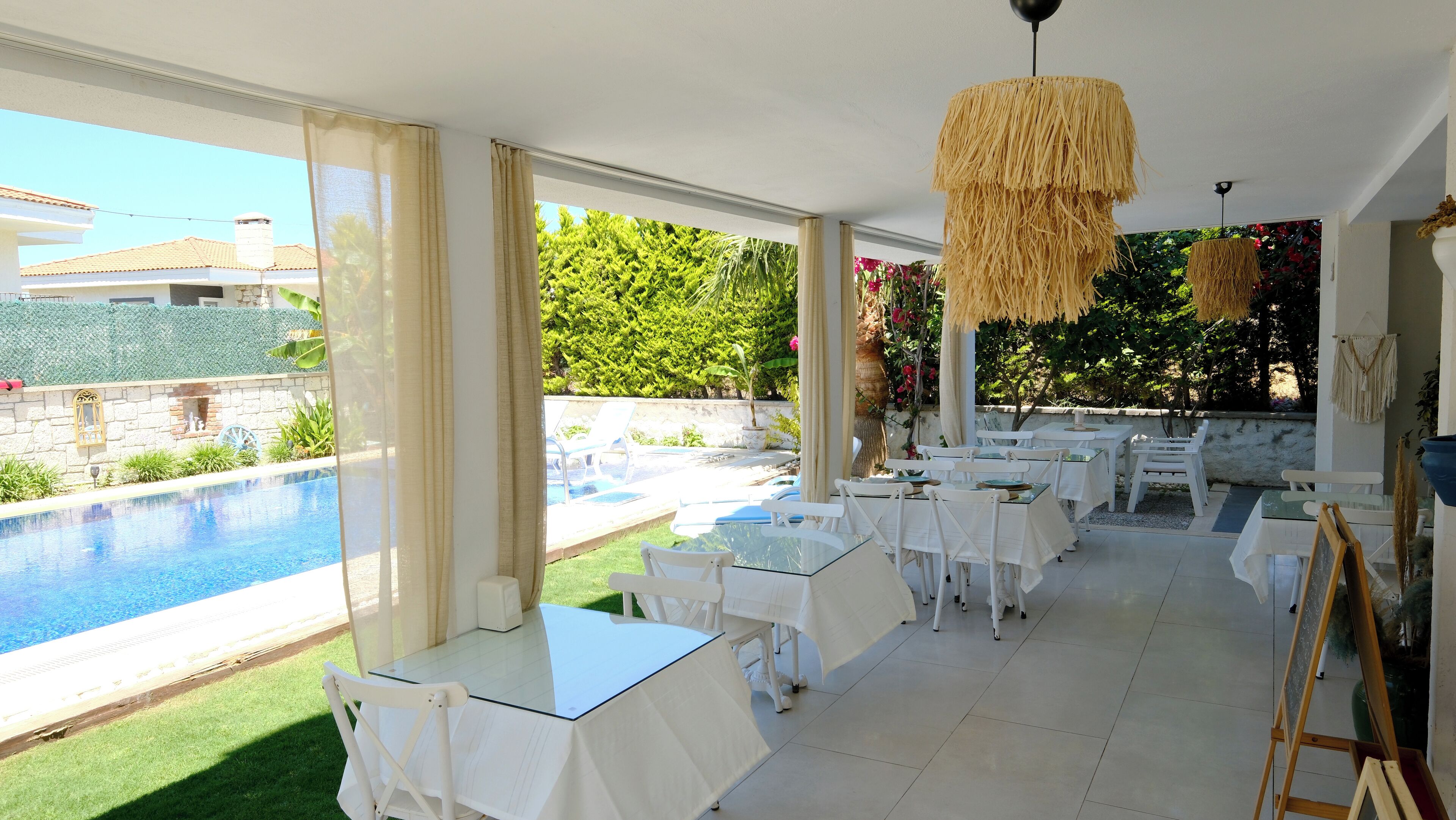 A sunlit outdoor dining area with a pool view, white tables and chairs, and a thatched ceiling light.