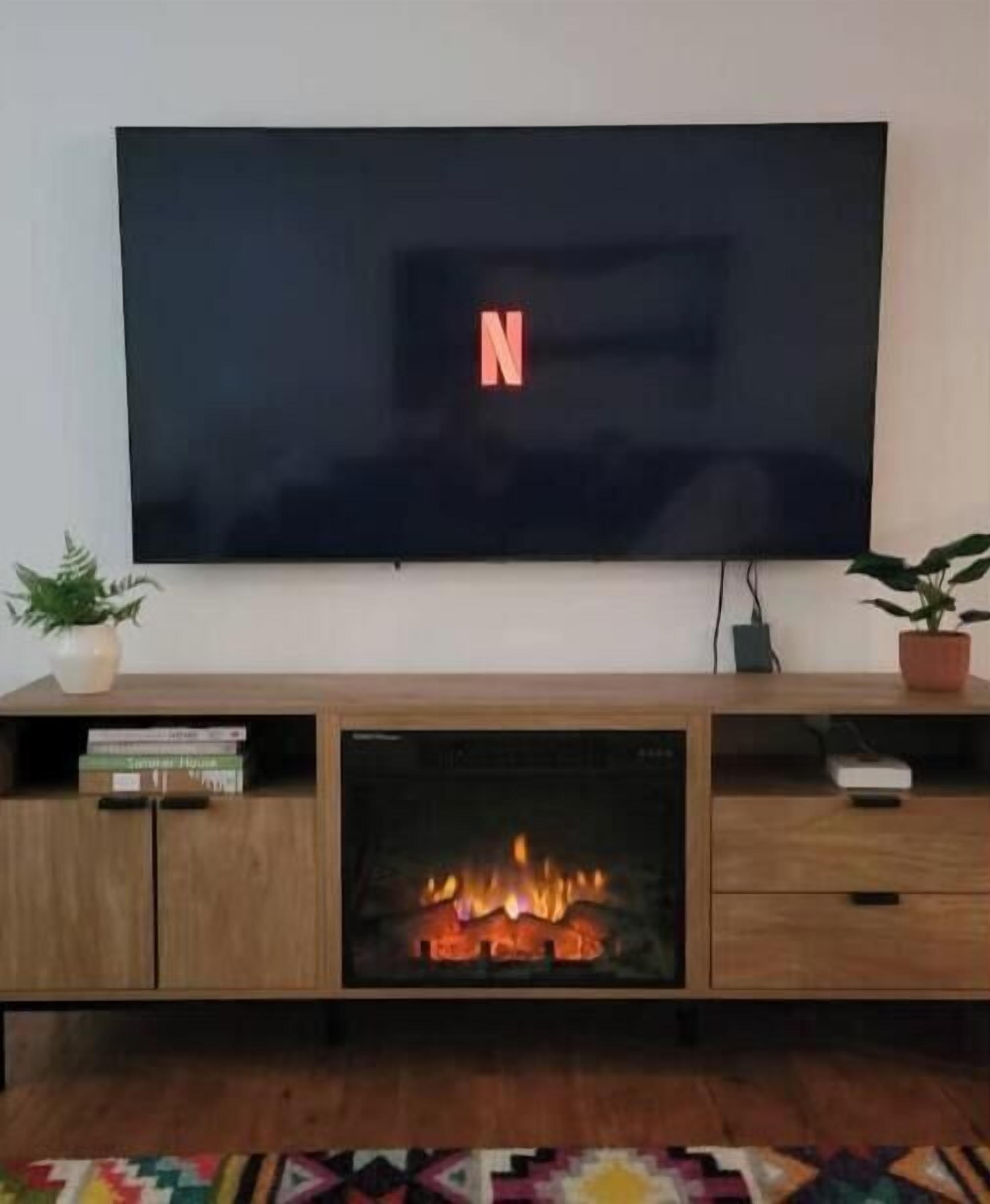 Una sala de estar moderna con un televisor de pantalla plana mostrando el logotipo de Netflix, una chimenea empotrada y un mueble de madera para entretenimiento con libros y objetos decorativos.