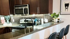 A modern kitchen with dark wooden cabinets, a stainless steel microwave, a gas stove, and a granite countertop. There are three black bar stools with wooden legs. - Lakeside Mountain Retreat - 3br, 2 bathroom beachfront condo with pool & hot tub (Invermere)