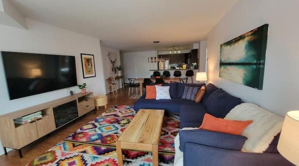 Een moderne woonkamer met een grote flatscreen televisie, een blauwe bank, een houten salontafel en een kleurrijk vloerkleed.