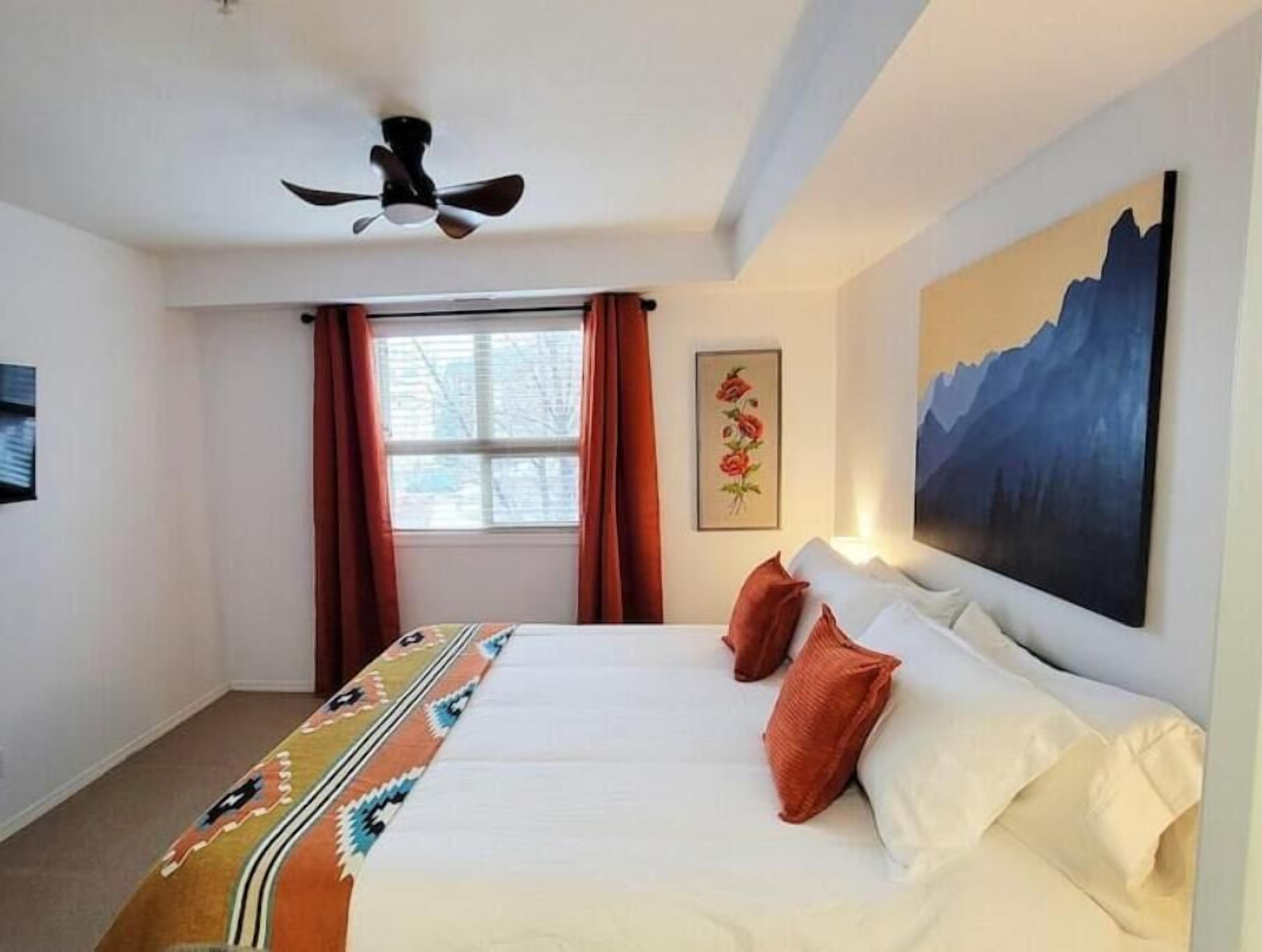 Een hotelkamer met een bed, een plafondventilator, een raam met gordijnen, een schilderij aan de muur en een vloerkleed met een geometrisch patroon.