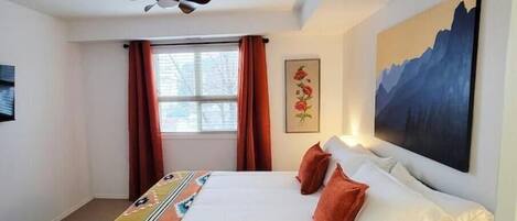 Een hotelkamer met een bed, een plafondventilator, een raam met gordijnen, een schilderij aan de muur en een vloerkleed met een geometrisch patroon.