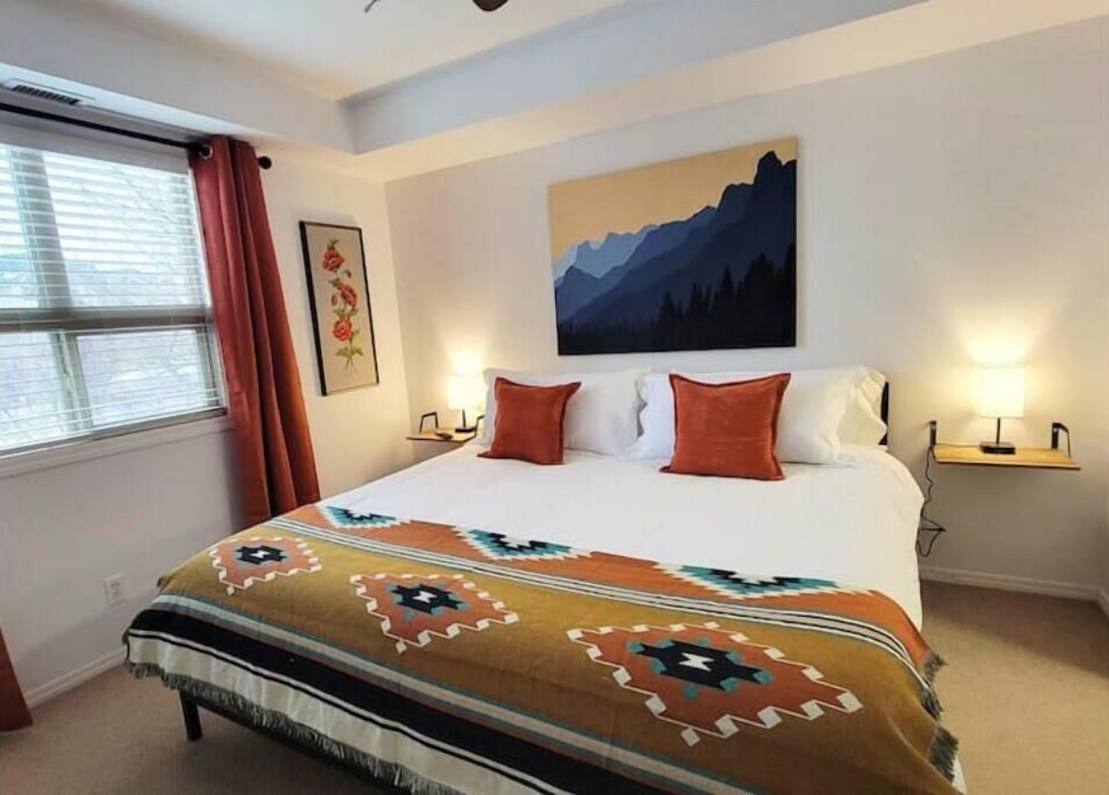A hotel room with a large bed, a mountain painting on the wall, a window with curtains, and a bedside table with a lamp.