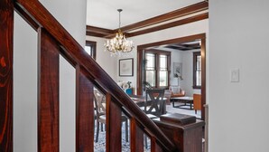 Una escalera de madera con un candelabro, que conduce a un salón con chimenea, una mesa de comedor y un sofá.