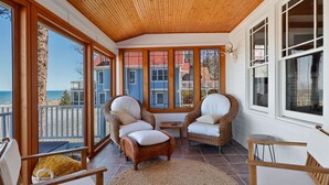 Una habitación luminosa con sillas de mimbre, una mesa de madera y grandes ventanales que dan a vistas de la playa y casas.