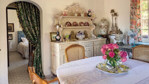 A room with a white ornate dresser, a table with a floral tablecloth, and a window with floral curtains. - VILLA LE MAS DE LA FORGE (Le Plan-de-la-Tour)