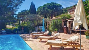 A poolside area with lounge chairs and umbrellas, surrounded by trees and a building with a tiled roof. - VILLA LE MAS DE LA FORGE (Le Plan-de-la-Tour)