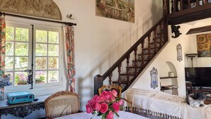 A room with a wooden staircase, a window with floral curtains, a table with a vase of flowers, and a painting on the wall. - VILLA LE MAS DE LA FORGE (Le Plan-de-la-Tour)