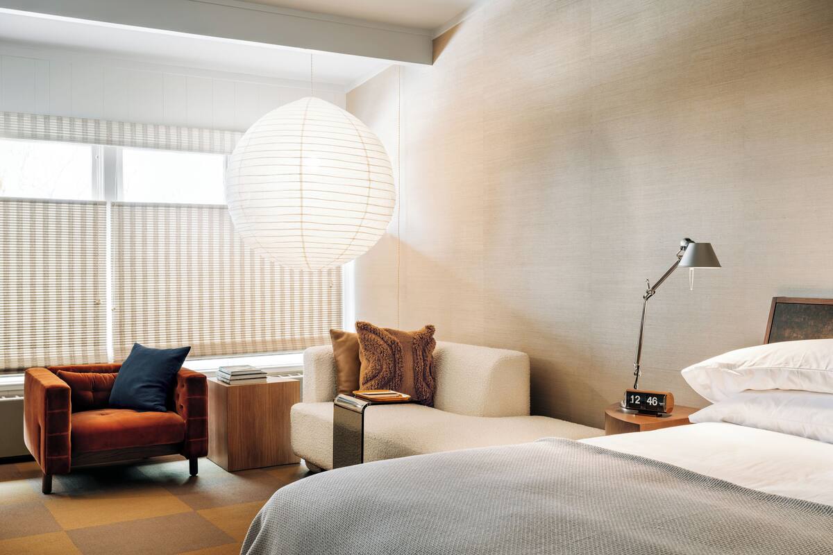A modern hotel room with a sofa, a chair, a bedside table with a lamp, and a large window with blinds.