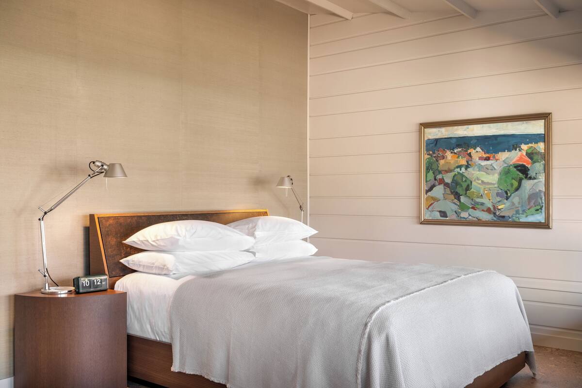 A neatly made bed with white linens, a wooden headboard, and two bedside lamps. A painting hangs on the wall to the right.
