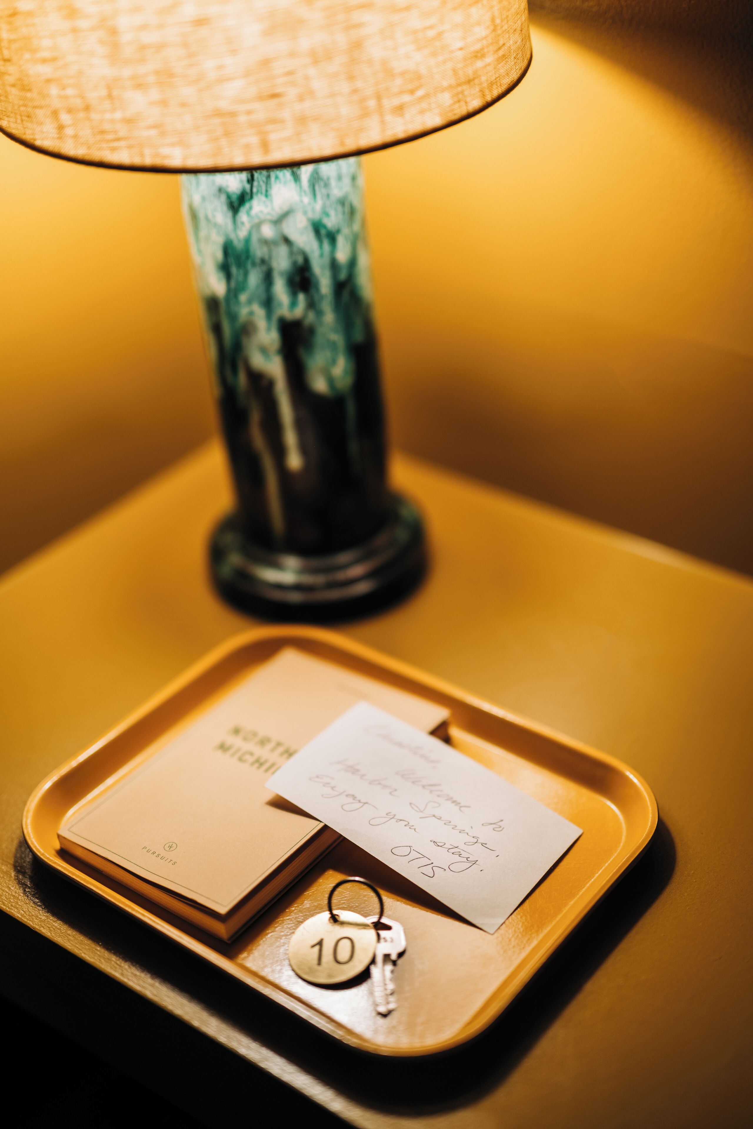 Un plateau contenant une clé de chambre d’hôtel, une carte et un petit guide, posé sur une table avec une lampe en arrière-plan.