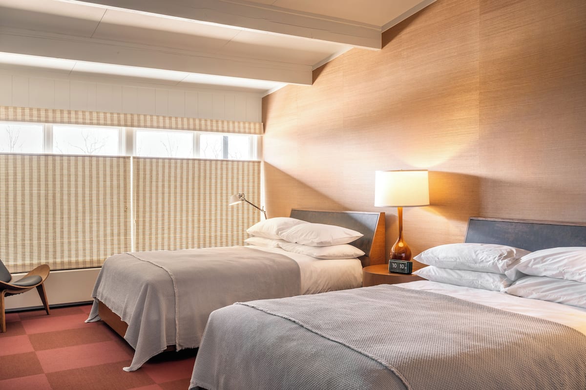 A hotel room with two beds, a nightstand with a lamp, a chair, and a window with blinds.