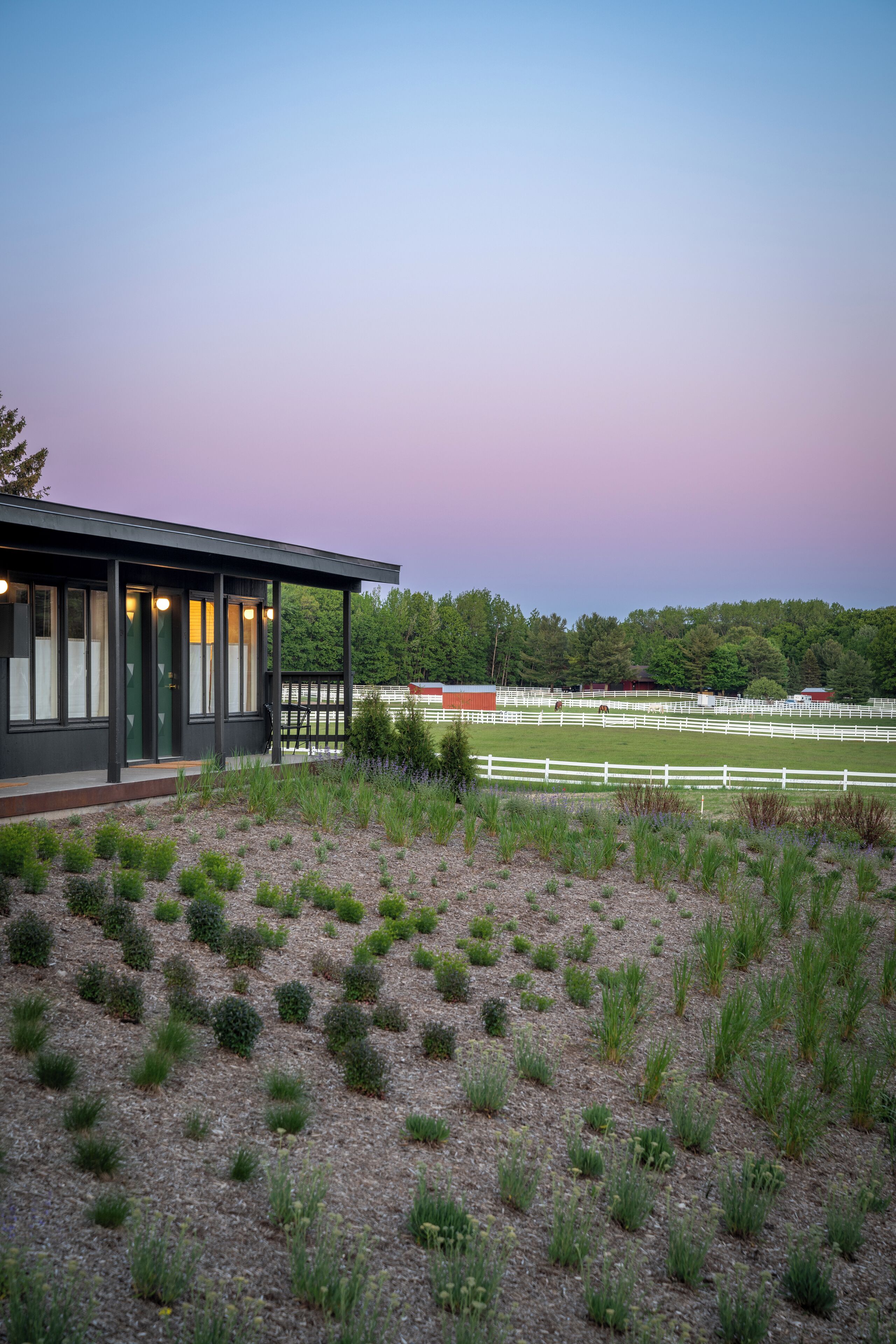 A modern building with a covered porch, surrounded by a fenced horse riding area and a natural landscape.