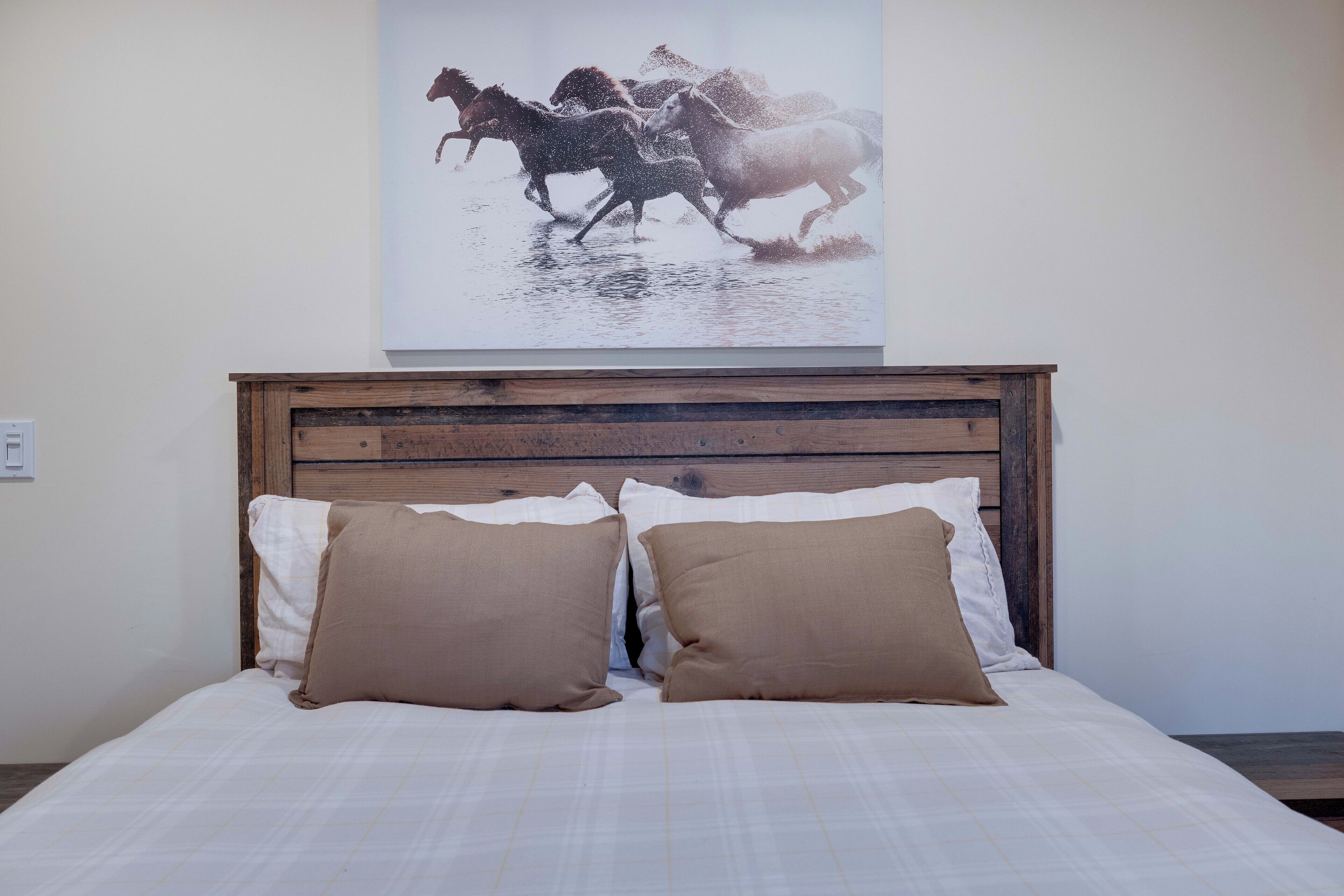 Un lit avec une tête de lit en bois, deux oreillers et une œuvre d’art représentant des chevaux, accrochée au mur.