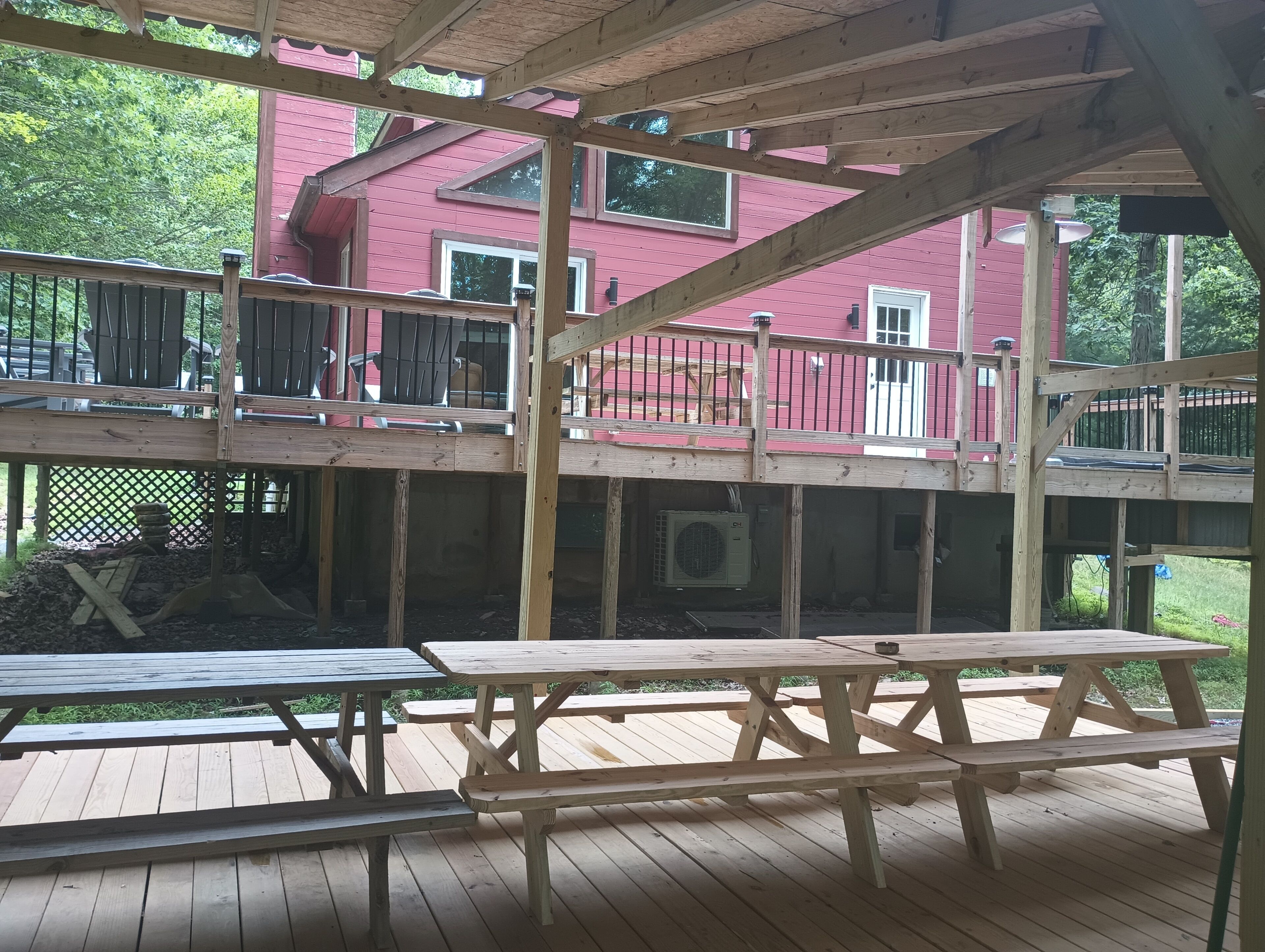 Une terrasse en bois avec des tables de pique-nique, un bâtiment rouge en arrière-plan et un espace abrité avec un toit en bois.