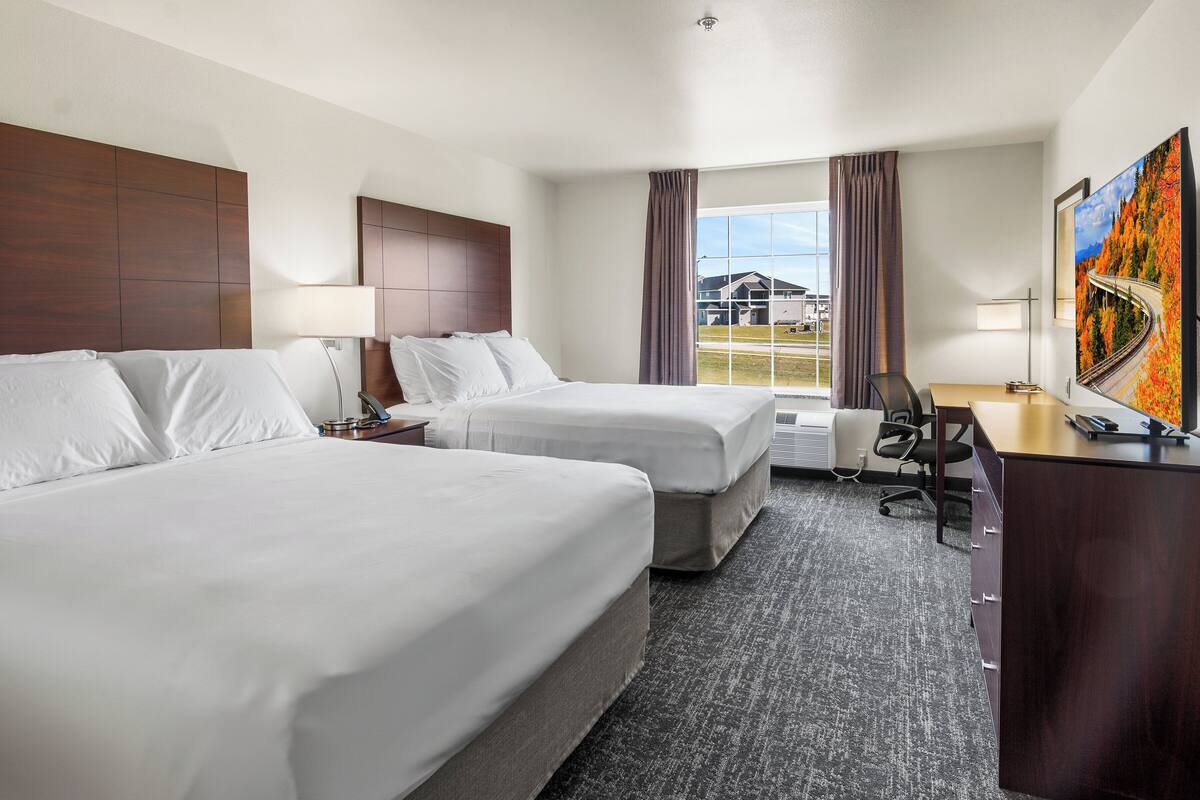 Quarto de hotel com duas camas, uma mesa de trabalho, uma cadeira e uma janela grande com cortinas.