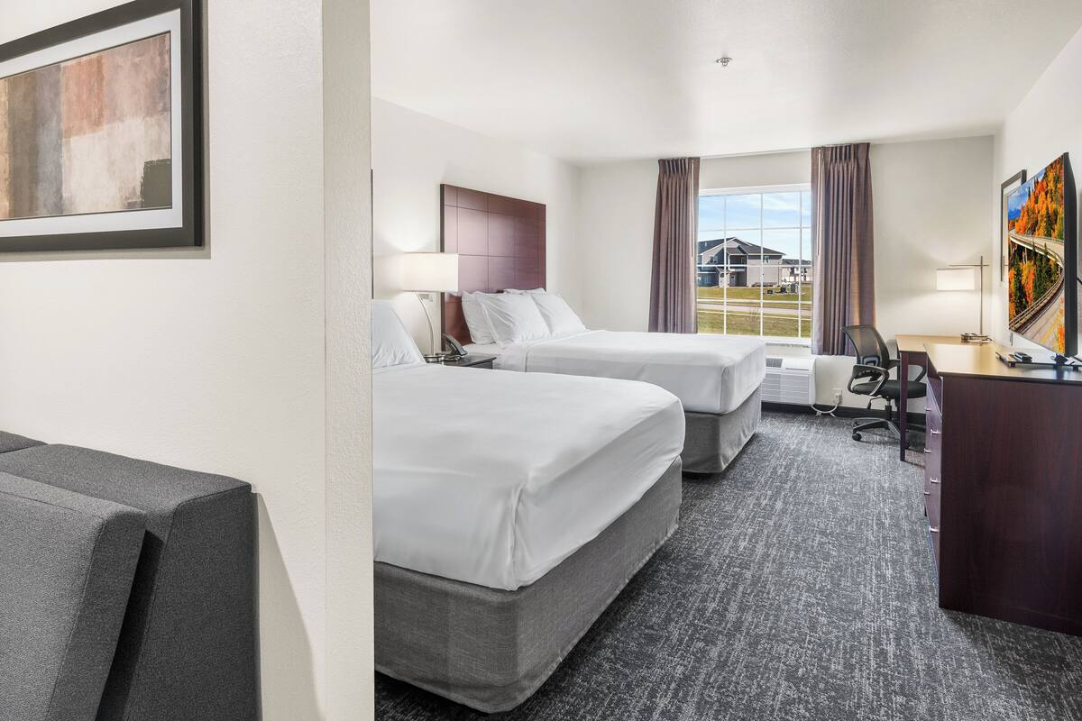 Quarto de hotel com duas camas, uma escrivaninha e uma cadeira. Há uma janela com vista para um edifício e um caminhão estacionado lá fora.
