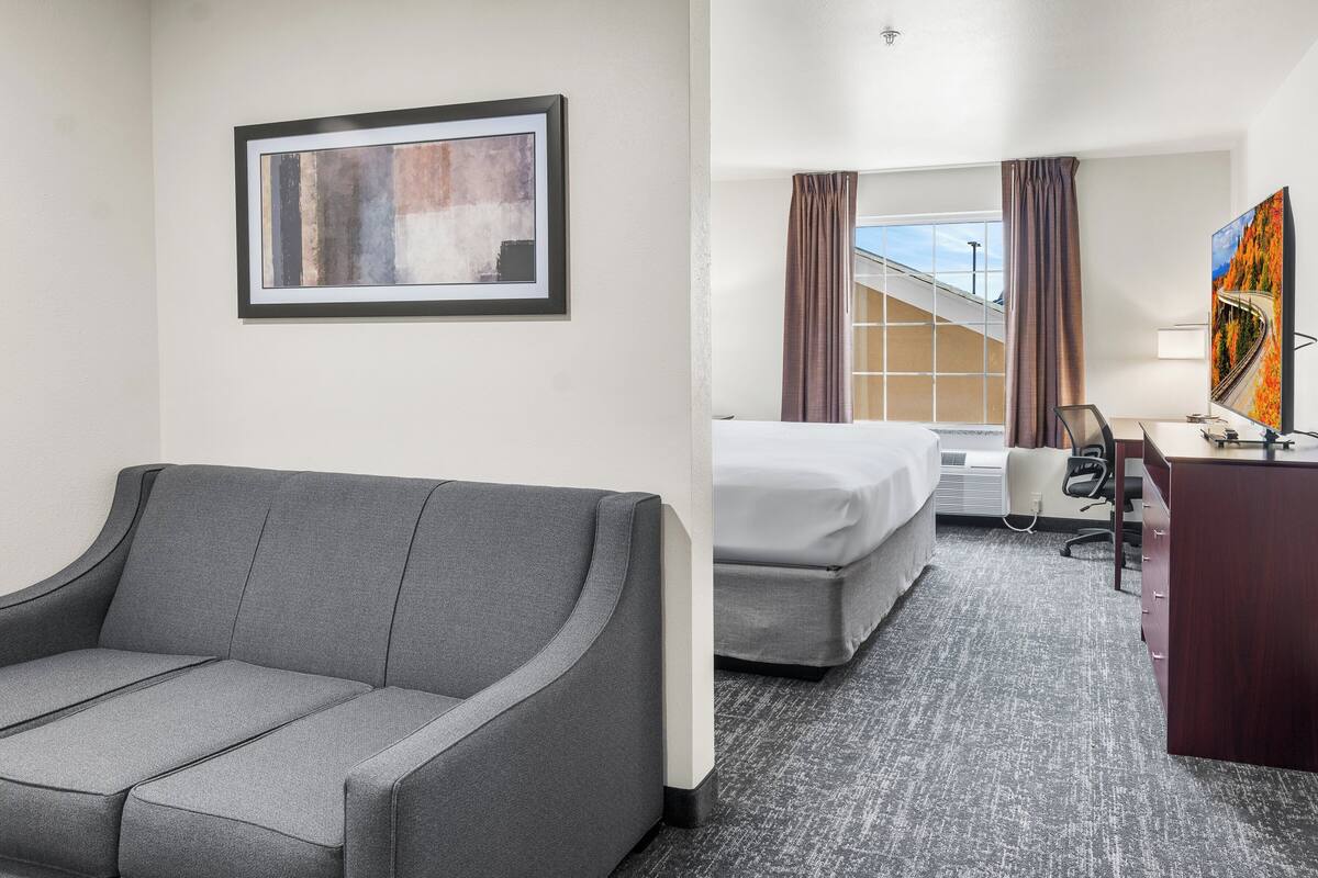 Quarto de hotel com sofá cinzento, cama, escrivaninha e uma janela grande com cortinas.
