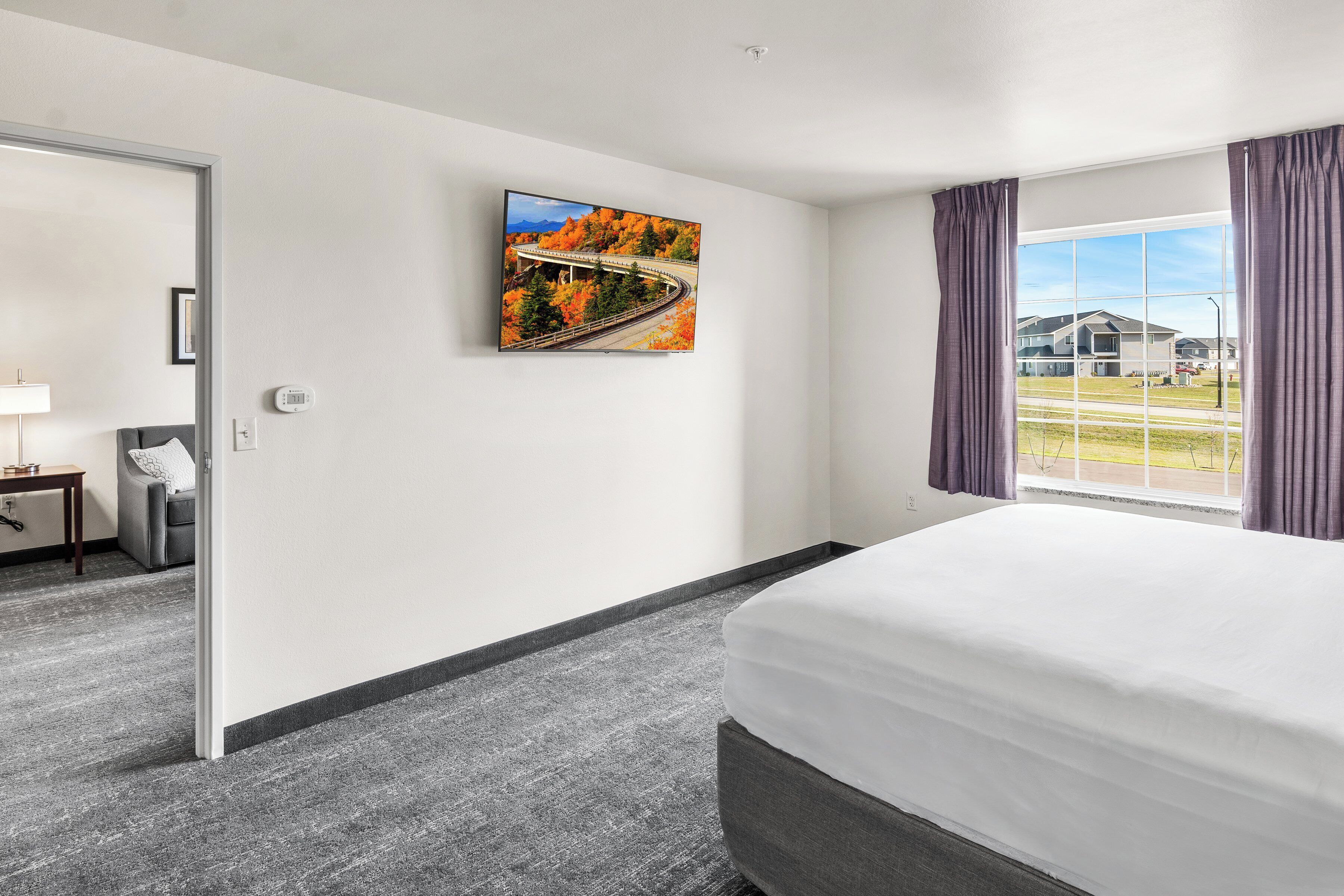 A hotel room with a bed, a painting of a bridge, a window with curtains, and a view of a building outside.