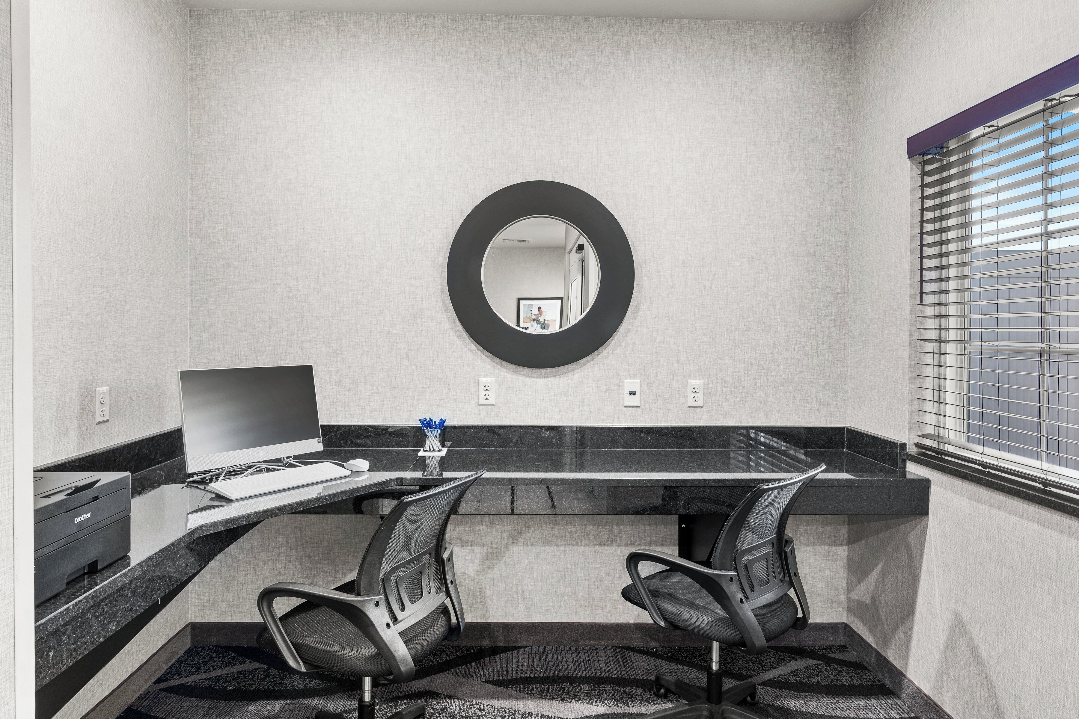 A modern office space with a large desk, two office chairs, a desktop computer, a printer, and a mirror on the wall.